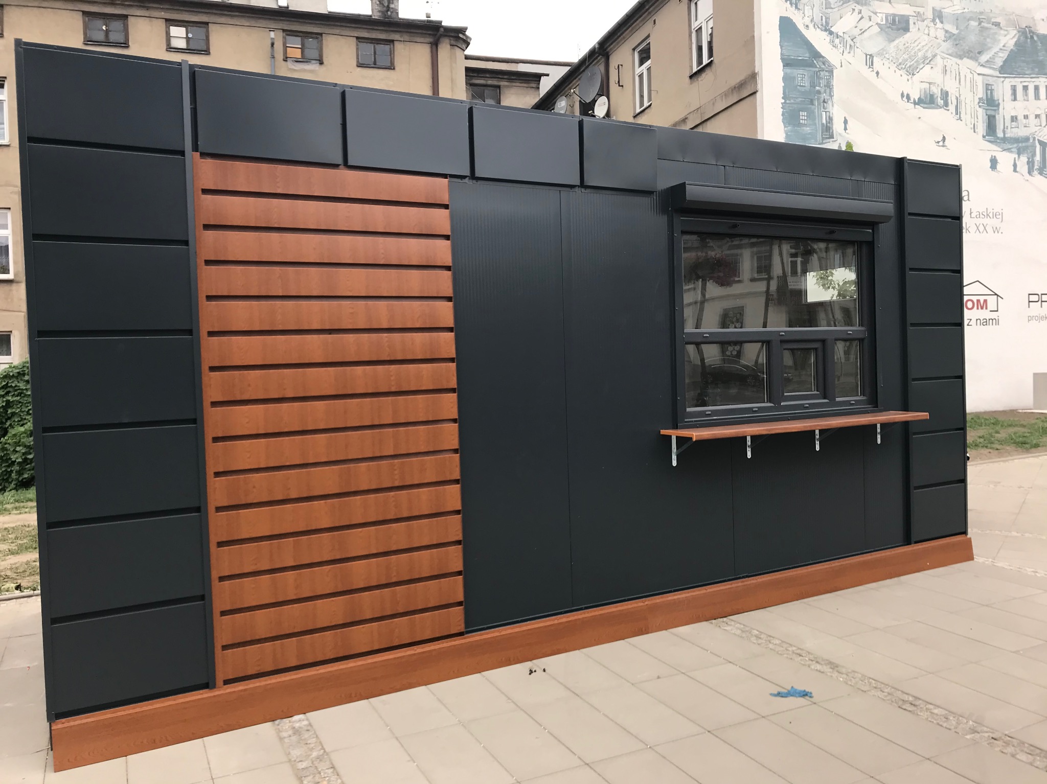 Nowoczesny pawilon z płyt warstwowych w kolorze grafitowym, z drewnianymi akcentami i oknem podawczym, na tle miejskiej zabudowy. Eleganckie wykończenie.