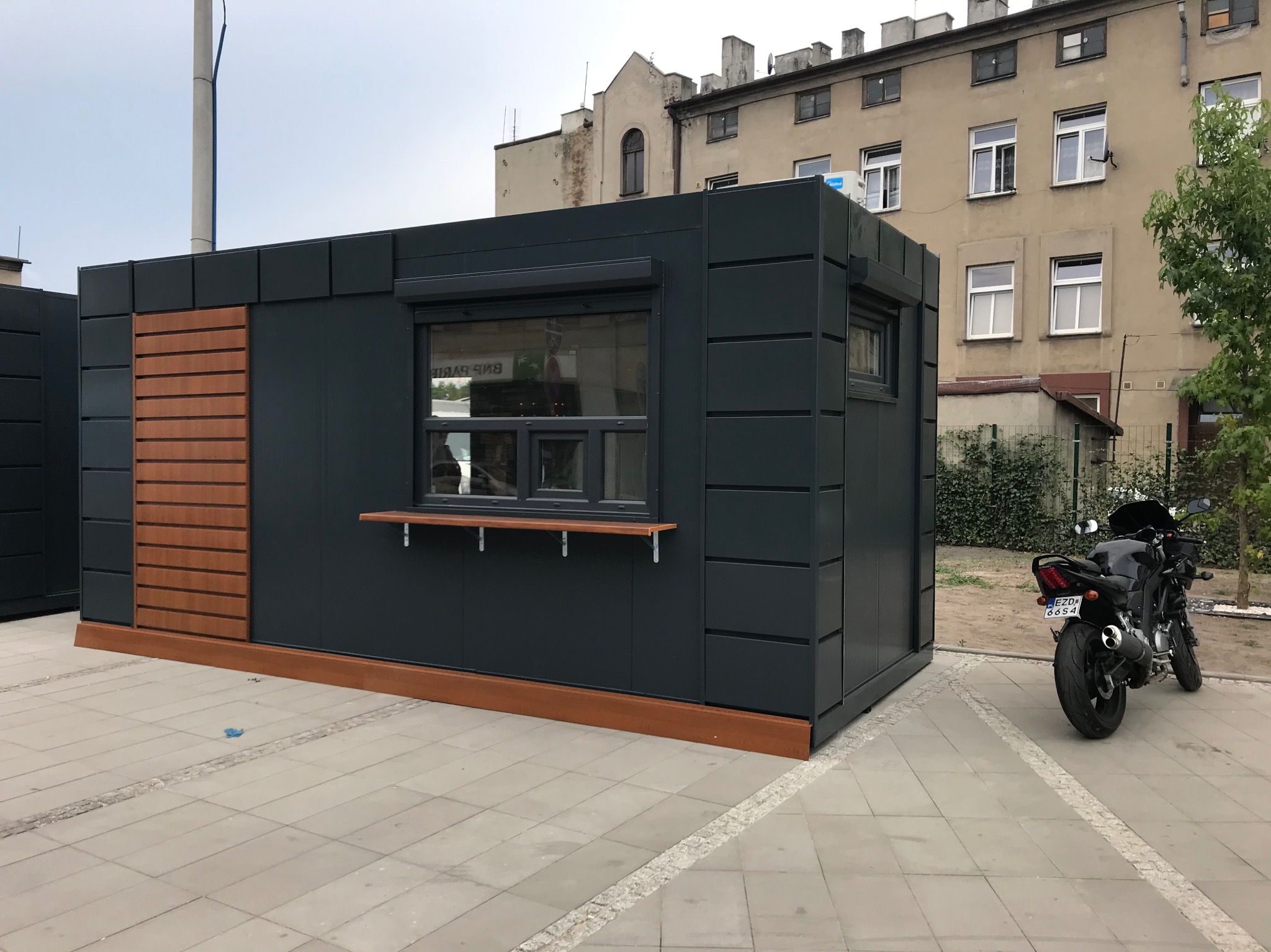 Gotowa, grafitowa budka handlowa z elementami drewnopodobnymi, z roletą i okienkiem podawczym, na tle budynku i zaparkowanego motocykla.