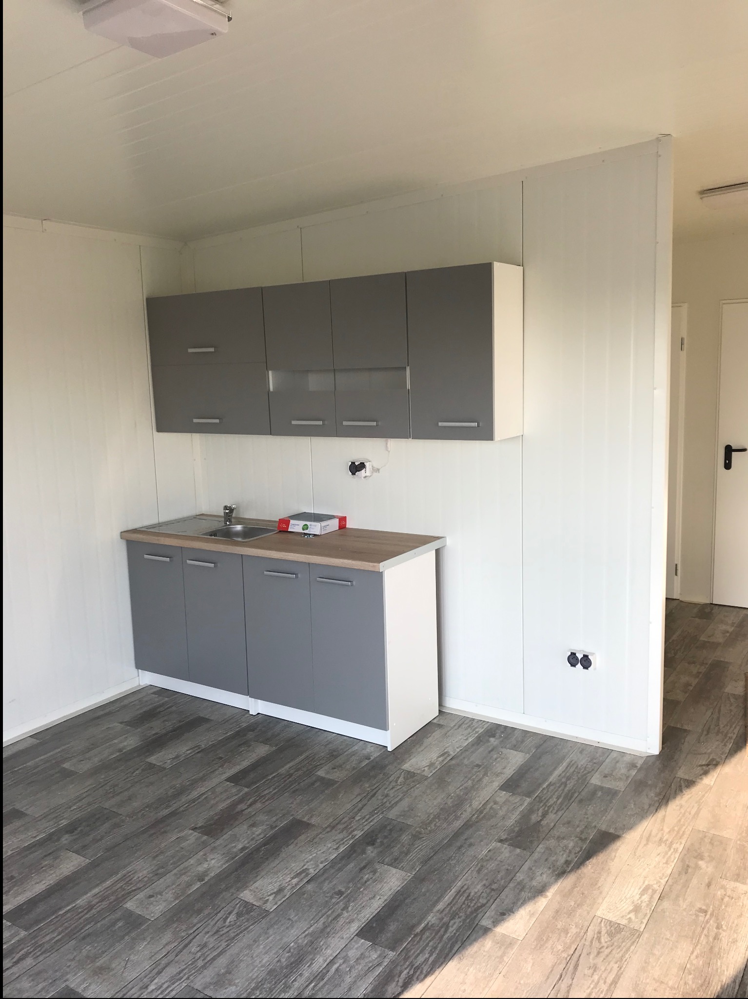 Wnętrze kontenera mieszkalnego z szarymi szafkami kuchennymi, białymi ścianami z płyt warstwowych i podłogą imitującą drewno. Widoczne gniazdka elektryczne.