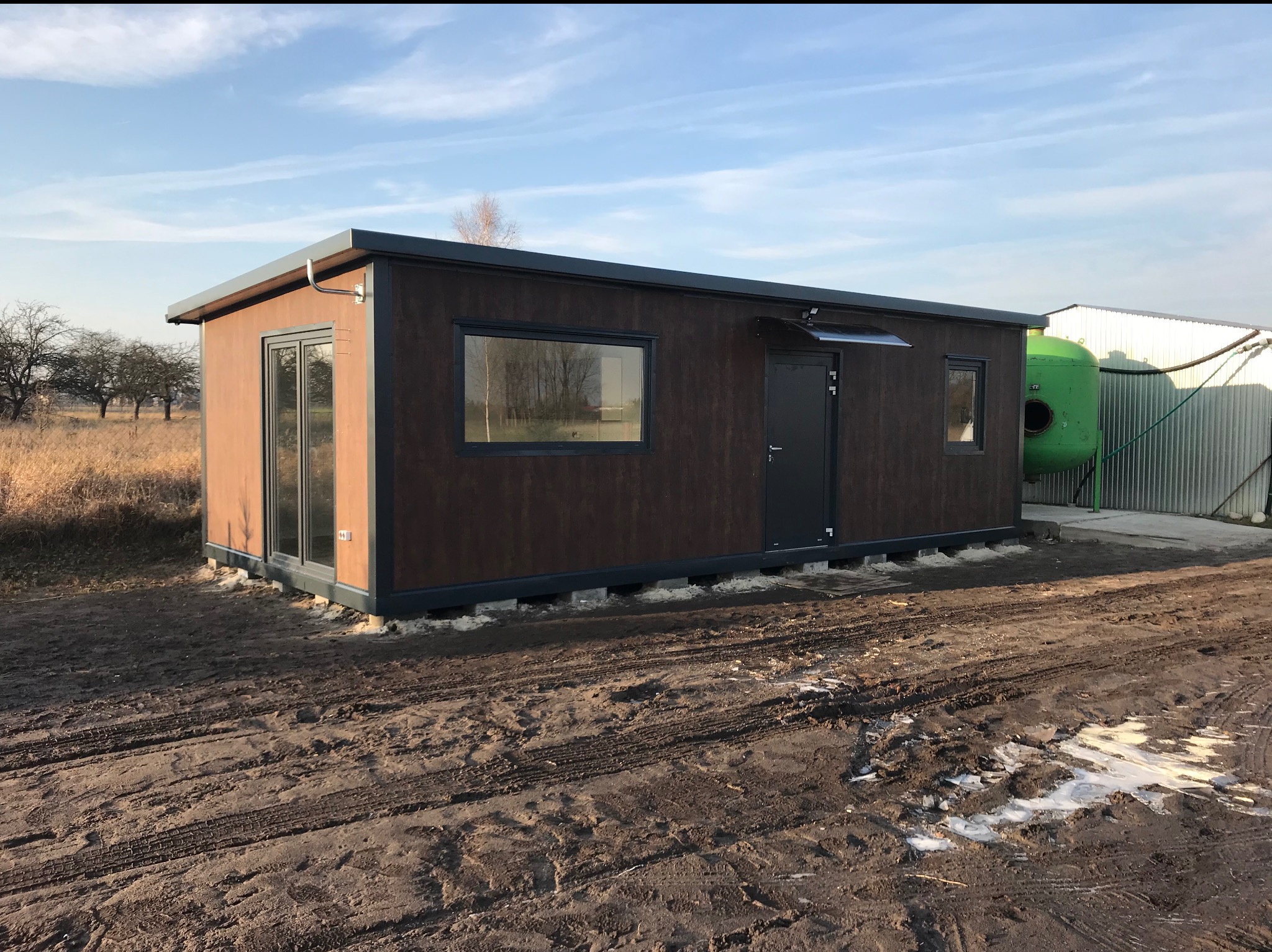 Gotowy, brązowy pawilon z płyt warstwowych z oknami i drzwiami, na nierównym terenie. W tle budynek z blachy falistej i zbiornik.