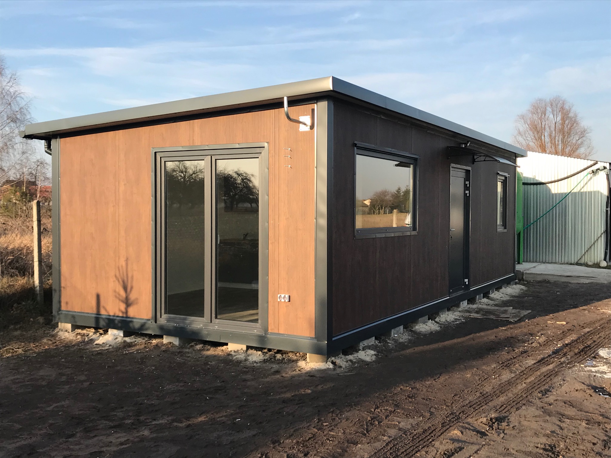 Gotowy pawilon z płyt warstwowych w brązowej kolorystyce, z oknami i drzwiami, posadowiony na betonowych bloczkach, w otoczeniu ziemi i częściowo widocznego ogrodzenia.