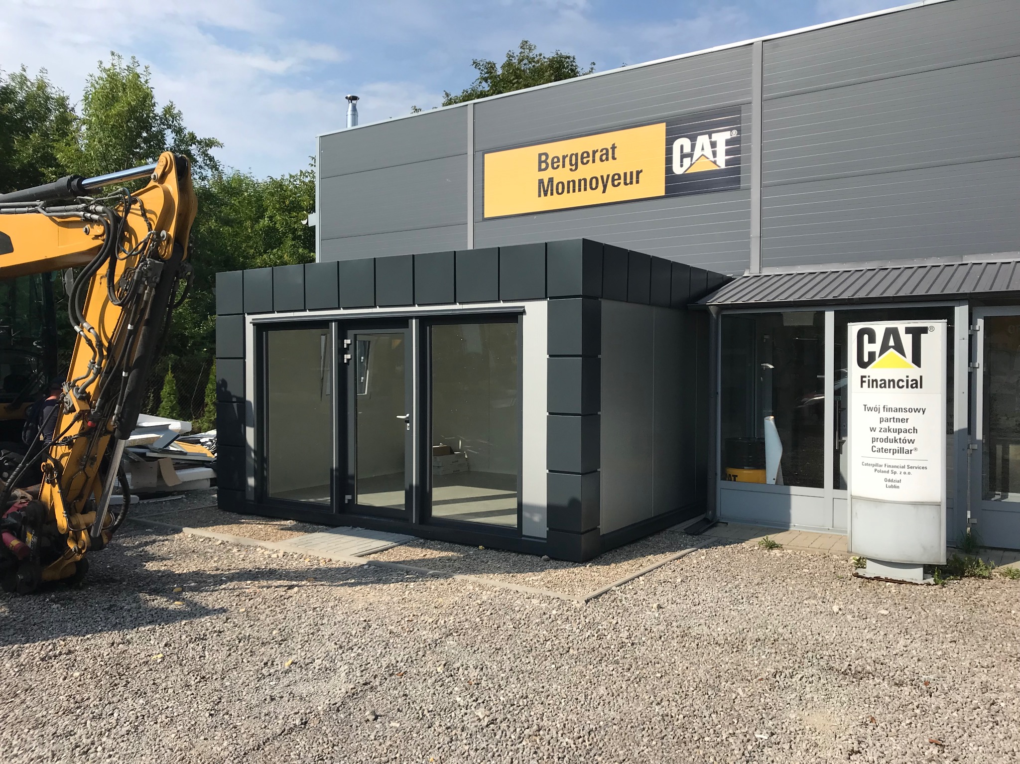 Szary budynek z płyt warstwowych z logo CAT i Bergerat Monnoyeur, częściowo zasłonięty przez żółtą koparkę. Widoczny napis CAT Financial na tablicy informacyjnej.