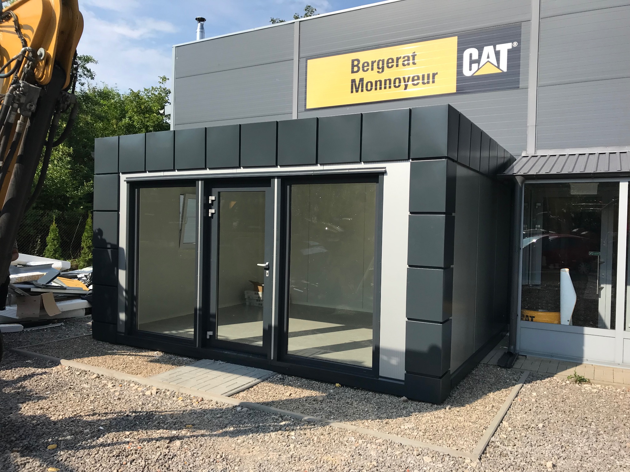 Nowoczesny pawilon biurowy z płyt warstwowych w szarych odcieniach, na tle budynku z logo CAT. Elementy koparki widoczne z lewej strony.