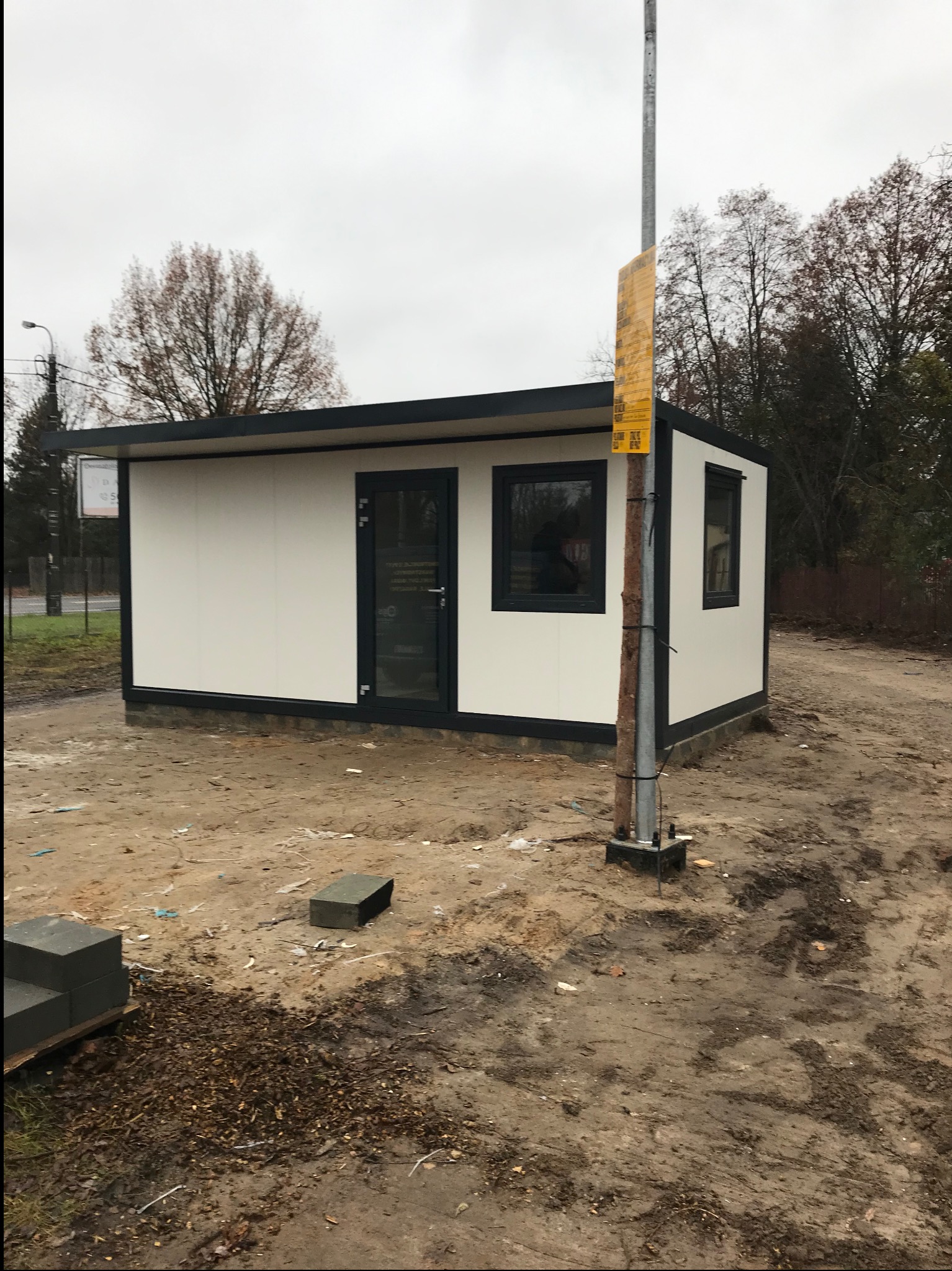 Biały pawilon z płyt warstwowych z czarnymi ramami okiennymi i drzwiami, stojący na placu budowy z widocznymi blokami i ziemią.