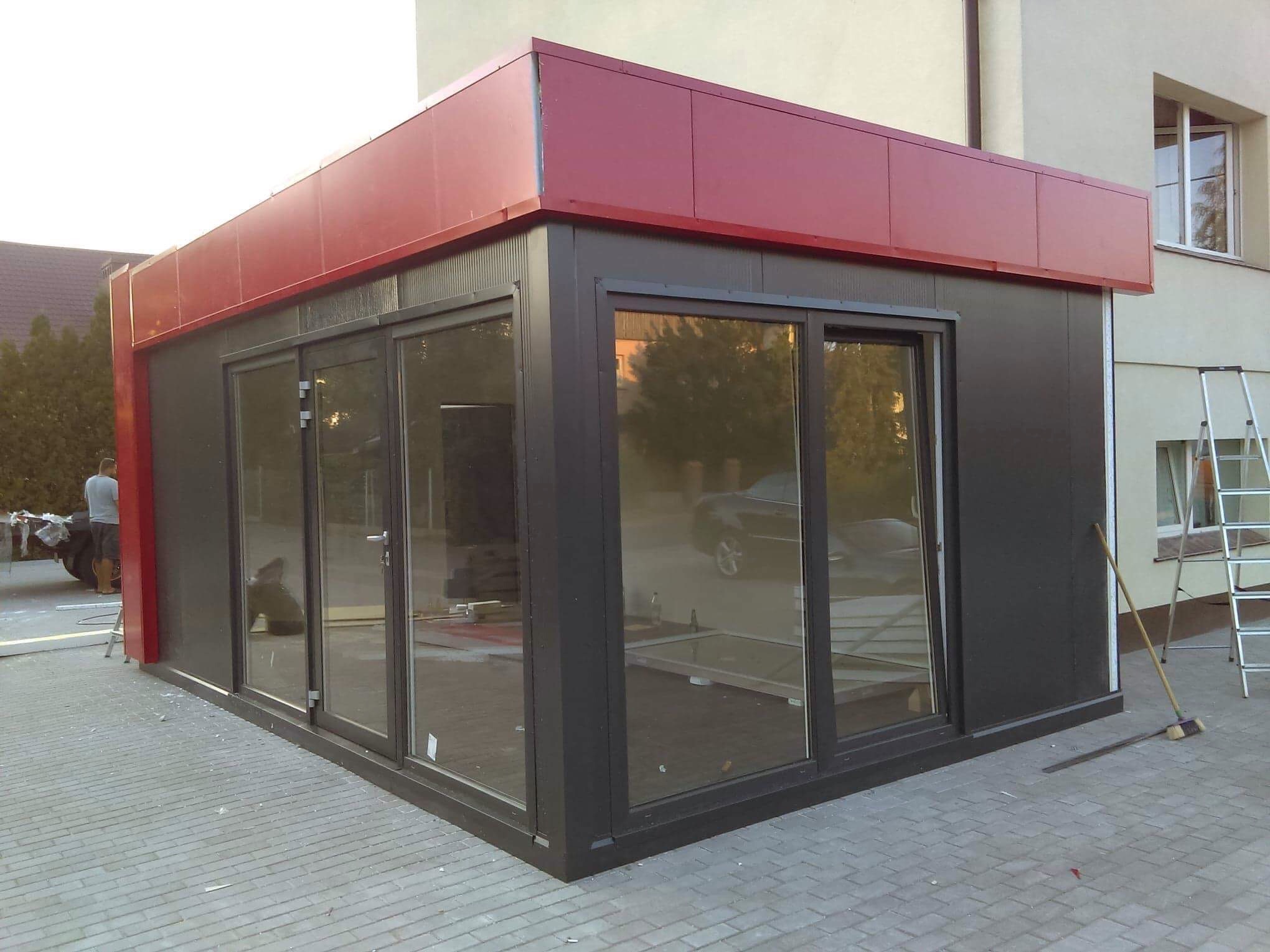 Pawilon z płyt warstwowych z dużymi oknami i czerwonym dachem, częściowo ukończony, widoczna drabina i narzędzia. Budynek w tle.