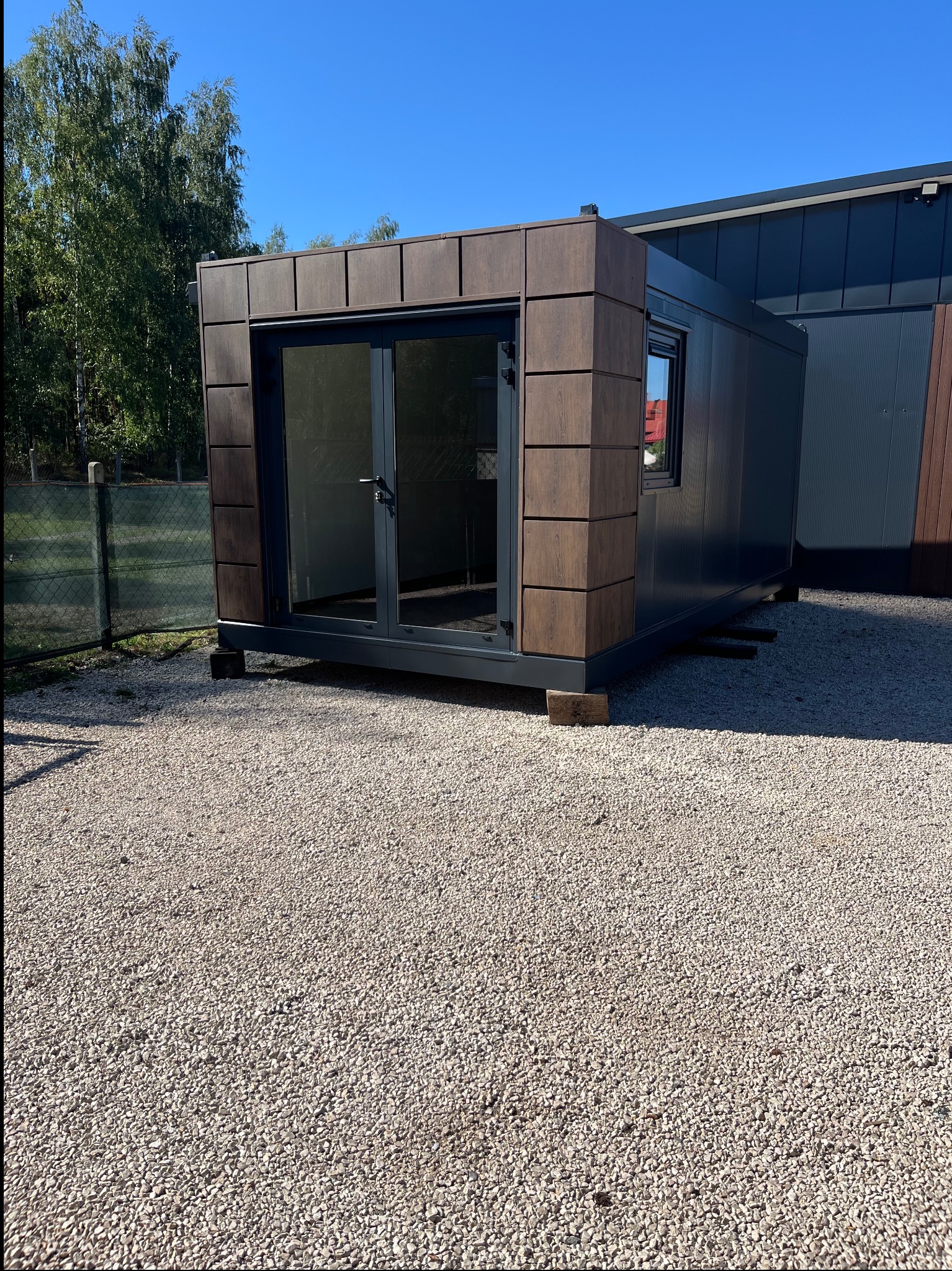 Gotowy modułowy budynek z płyt warstwowych z oknami i szklanymi drzwiami na żwirowym podłożu, widok z zewnątrz w słoneczny dzień.