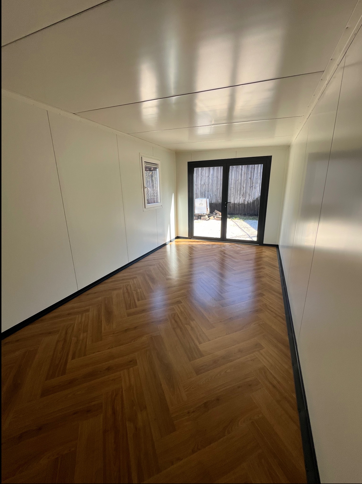 Wnętrze kontenera z płyt warstwowych: parkiet w jodełkę, białe ściany, okno i przeszklone drzwi z widokiem na zewnątrz. Jasne, słoneczne światło.