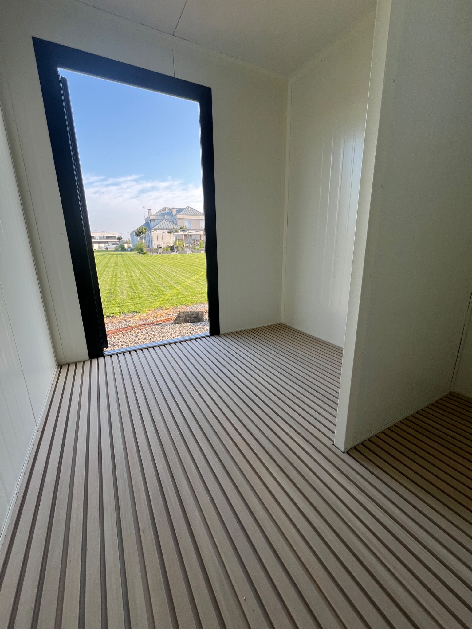 Wnętrze kontenera z panelami ściennymi i podłogą z desek, widok na zewnątrz przez czarną ramę okienną na trawnik i budynek w tle.