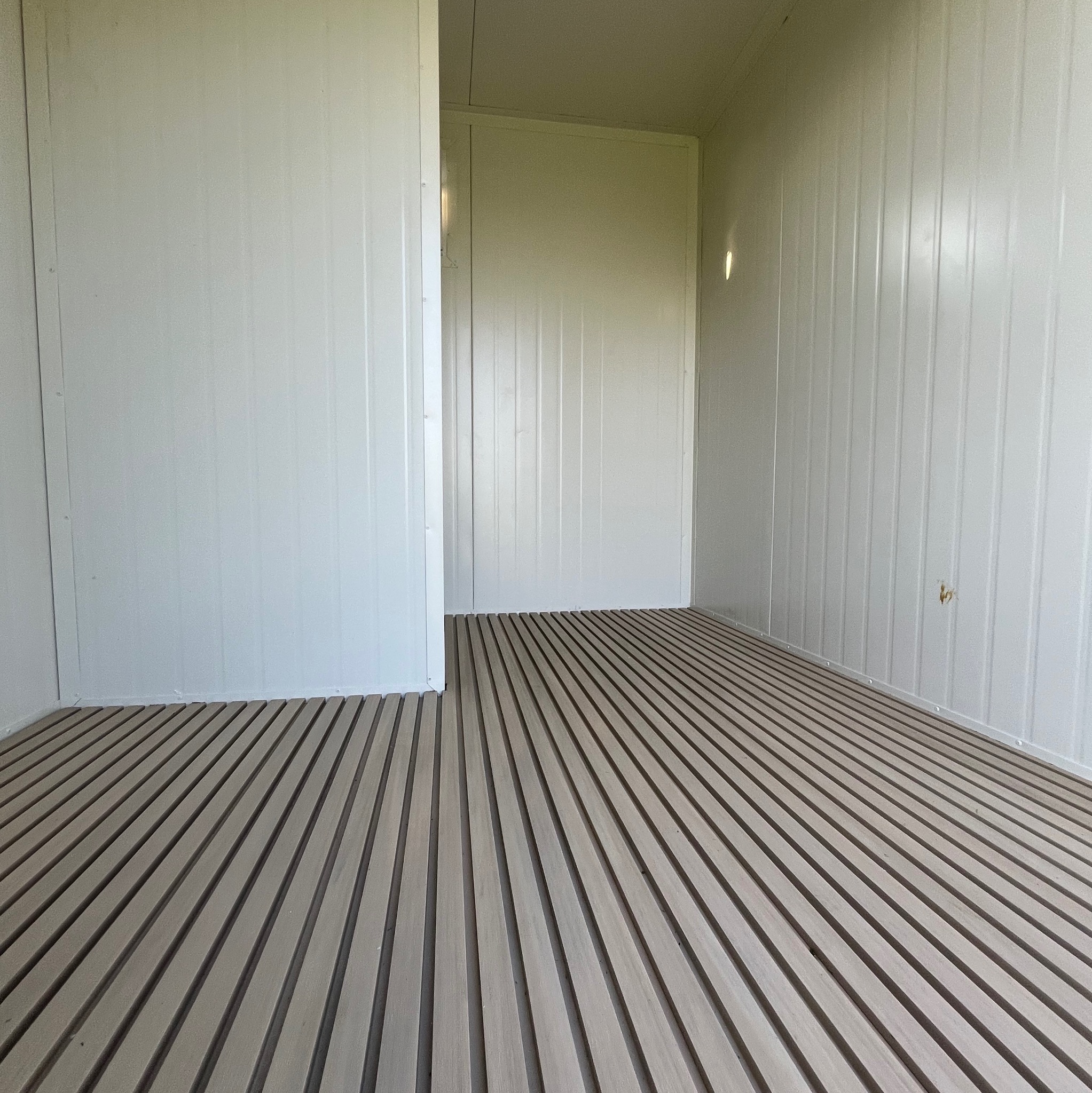 Wnętrze hali z płyt warstwowych: białe ściany z pionowymi przetłoczeniami i podłoga z drewnianych listew ułożonych równolegle. Widok na sąsiednie pomieszczenie.