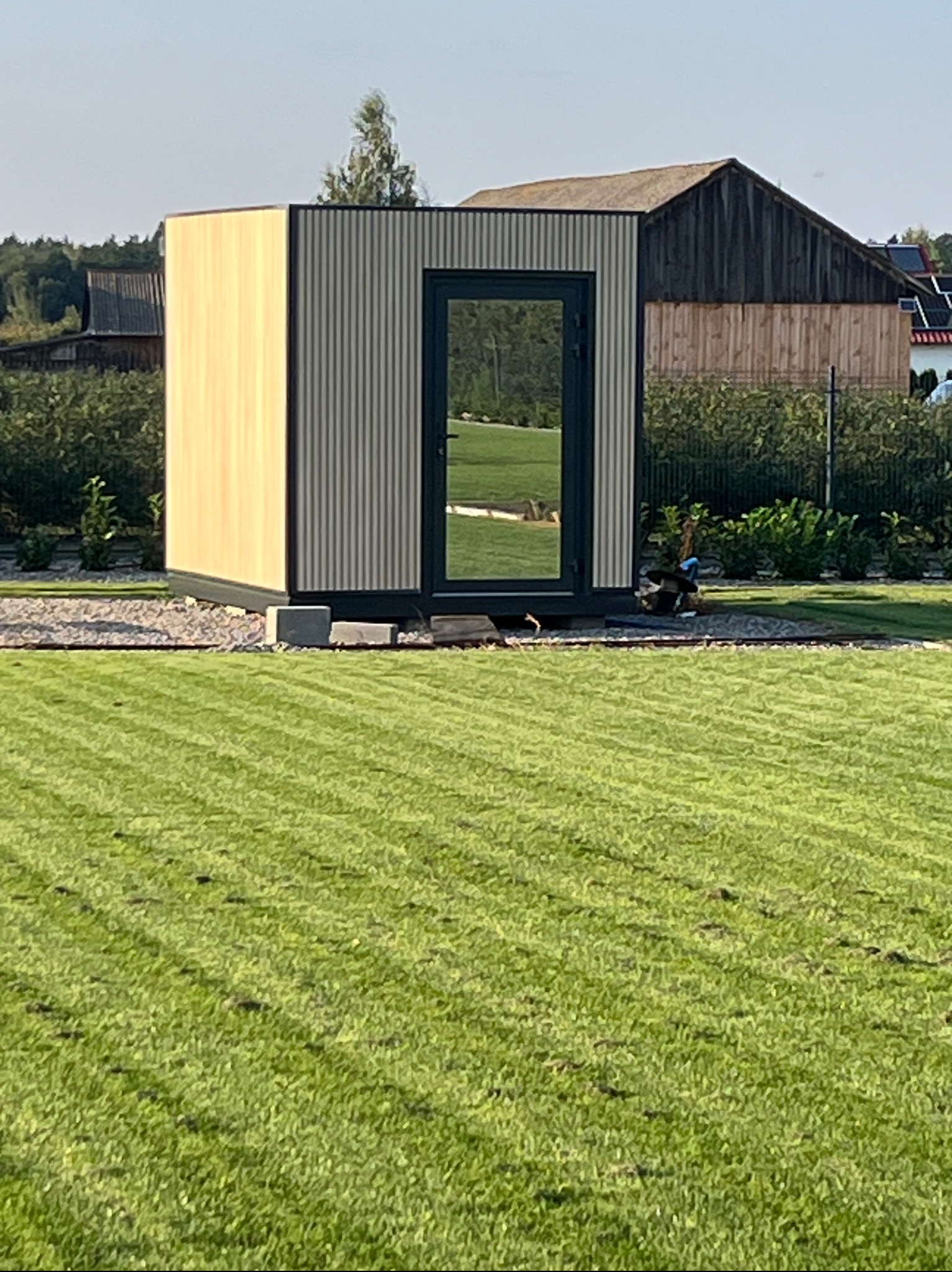 Nowoczesny, modułowy domek ogrodowy z płyt warstwowych, z czarną stolarką, umieszczony na trawniku. Widoczne elementy konstrukcji i otoczenie.