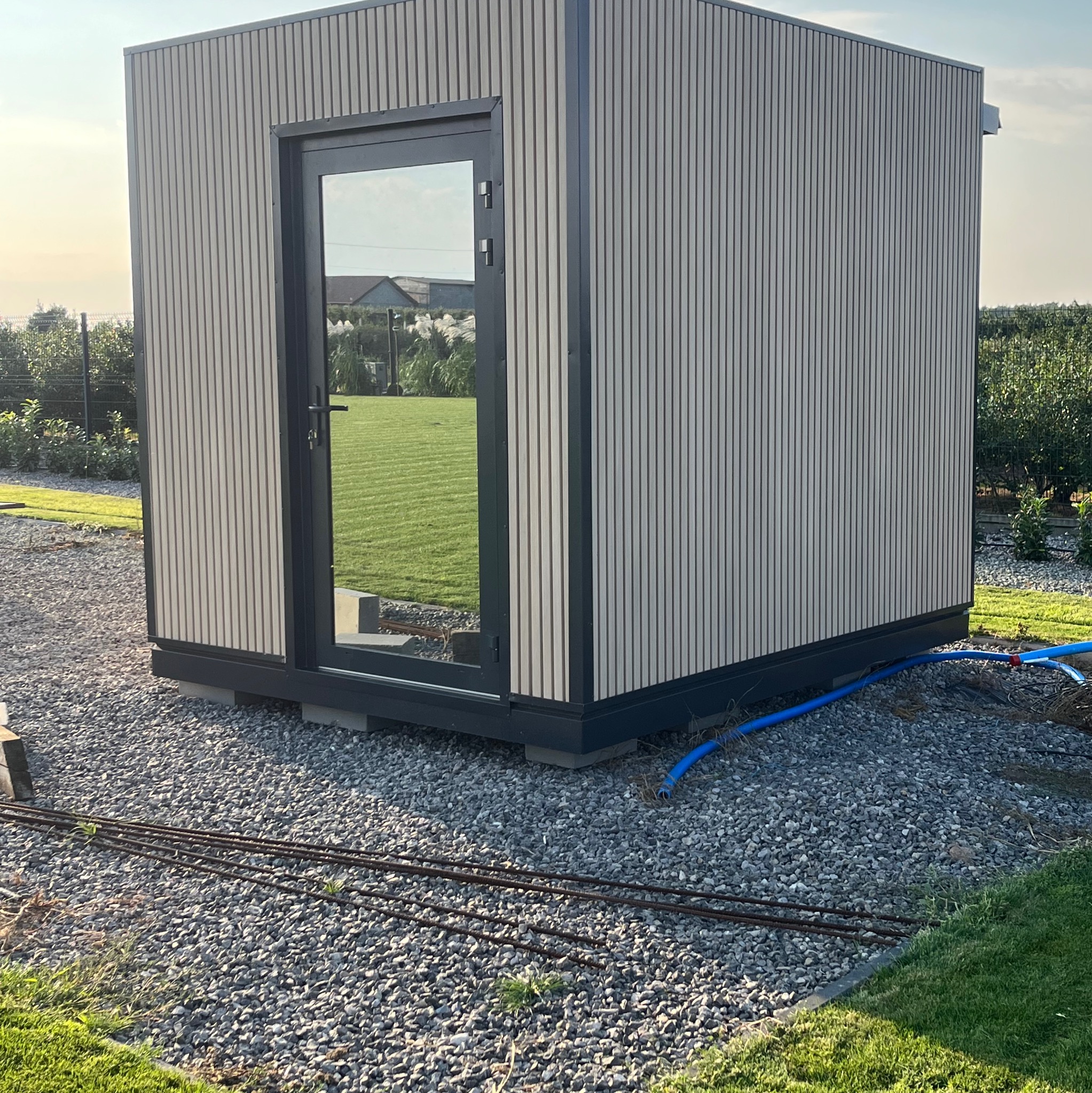 Nowoczesna, modułowa konstrukcja z jasnych paneli warstwowych i ciemną stolarką okienną. Całość osadzona na żwirowym podłożu z widocznymi elementami instalacji.