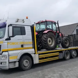 Ciężarówka pomocy drogowej MAN z załadowanym czerwonym traktorem rolniczym na platformie, w tle widoczne inne traktory i budynki gospodarcze.