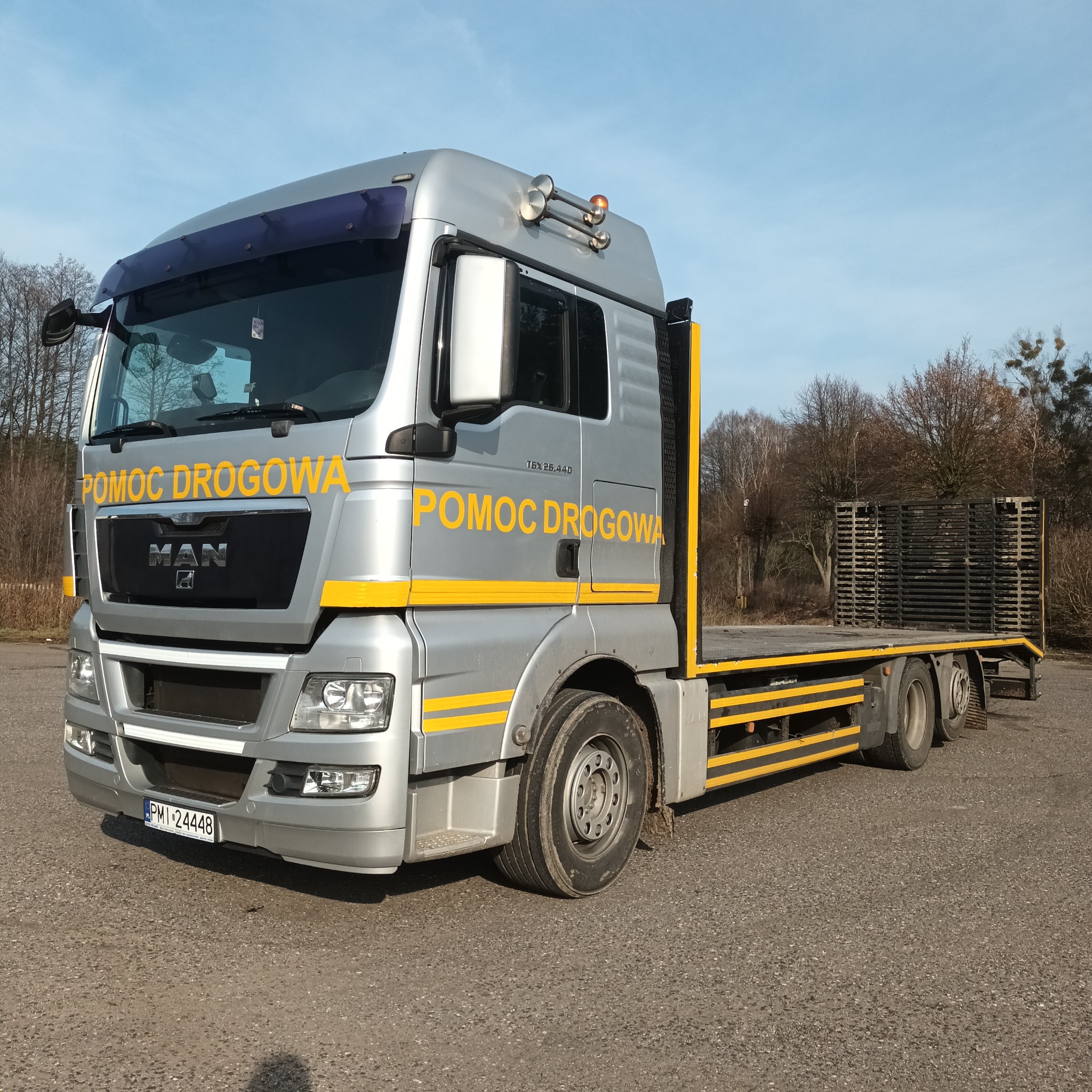 Srebrna ciężarówka z platformą i napisem 'Pomoc Drogowa' na tle szarego parkingu i drzew bez liści pod błękitnym niebem.