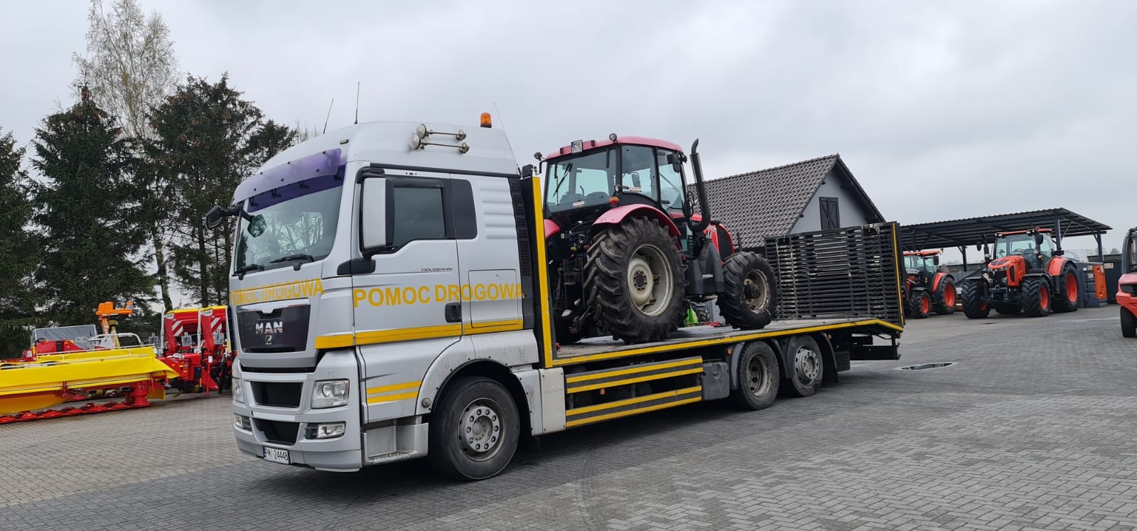 Ciężarówka pomocy drogowej MAN z załadowanym czerwonym traktorem rolniczym na platformie, w tle widoczne inne traktory i budynki gospodarcze.