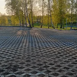 Ułożona nawierzchnia z ażurowych płyt betonowych na podbudowie piaskowej, z widocznym fragmentem ogrodzenia panelowego i zielenią w tle, oświetlona popołudniowym słońcem.