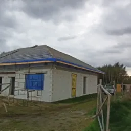 Surowy stan budynku jednorodzinnego z pustaków, z częściowo ułożonym dachem pokrytym szarą dachówką, zabezpieczonym niebieską folią, na tle pochmurnego nieba i zalesionego terenu.