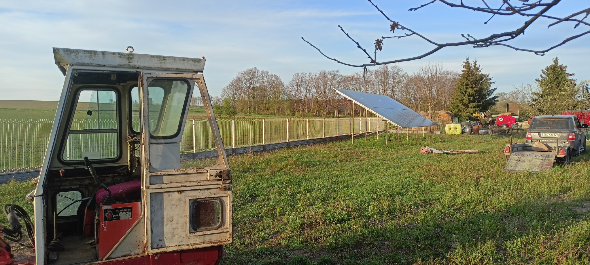 Widok z kabiny starego mini-koparki na ogrodzenie z siatki, panele słoneczne i zaparkowany samochód z przyczepą w tle krajobrazu.