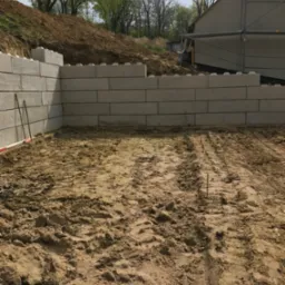 Wzniesiony mur oporowy z betonowych bloków w kształcie klocków, widoczne ślady opon na ziemi, w tle zbocze i budynek w trakcie budowy.
