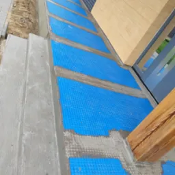 Hydroizolacja fundamentów budynku z użyciem folii kubełkowej, widoczne betonowe schody wejściowe i drewniany słup podtrzymujący zadaszenie.