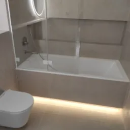 Nowoczesna łazienka w odcieniach szarości z podświetlaną wanną za szklanym panelem i podwieszaną toaletą.