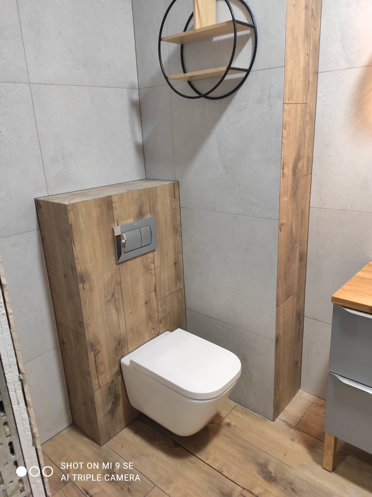 Nowoczesna toaleta podwieszana z zabudową z paneli drewnopodobnych, szare płytki ceramiczne na ścianach, okrągły regał wiszący z drewnianymi półkami, drewniana podłoga.