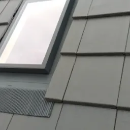 Szare dachówki ceramiczne z zamontowanym oknem dachowym z ciemną obwódką i specjalną uszczelniającą listwą pod oknem.