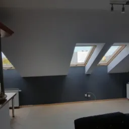 Poddasze z trzema oknami dachowymi w białych ramach, widok na zewnątrz, ciemnoszare ściany, jasna podłoga, widoczny fragment baru z wyposażeniem.
