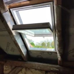 Częściowo otwarte okno dachowe, widok z wnętrza poddasza, z widoczną konstrukcją drewnianą i izolacją, z zewnątrz widok na zieleń.