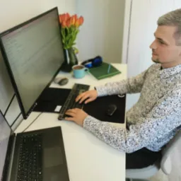 Mężczyzna w kwiecistej koszuli siedzi przy biurku z dwoma monitorami i laptopem, analizując wykresy, obok wazon z tulipanami i niebieski kubek.