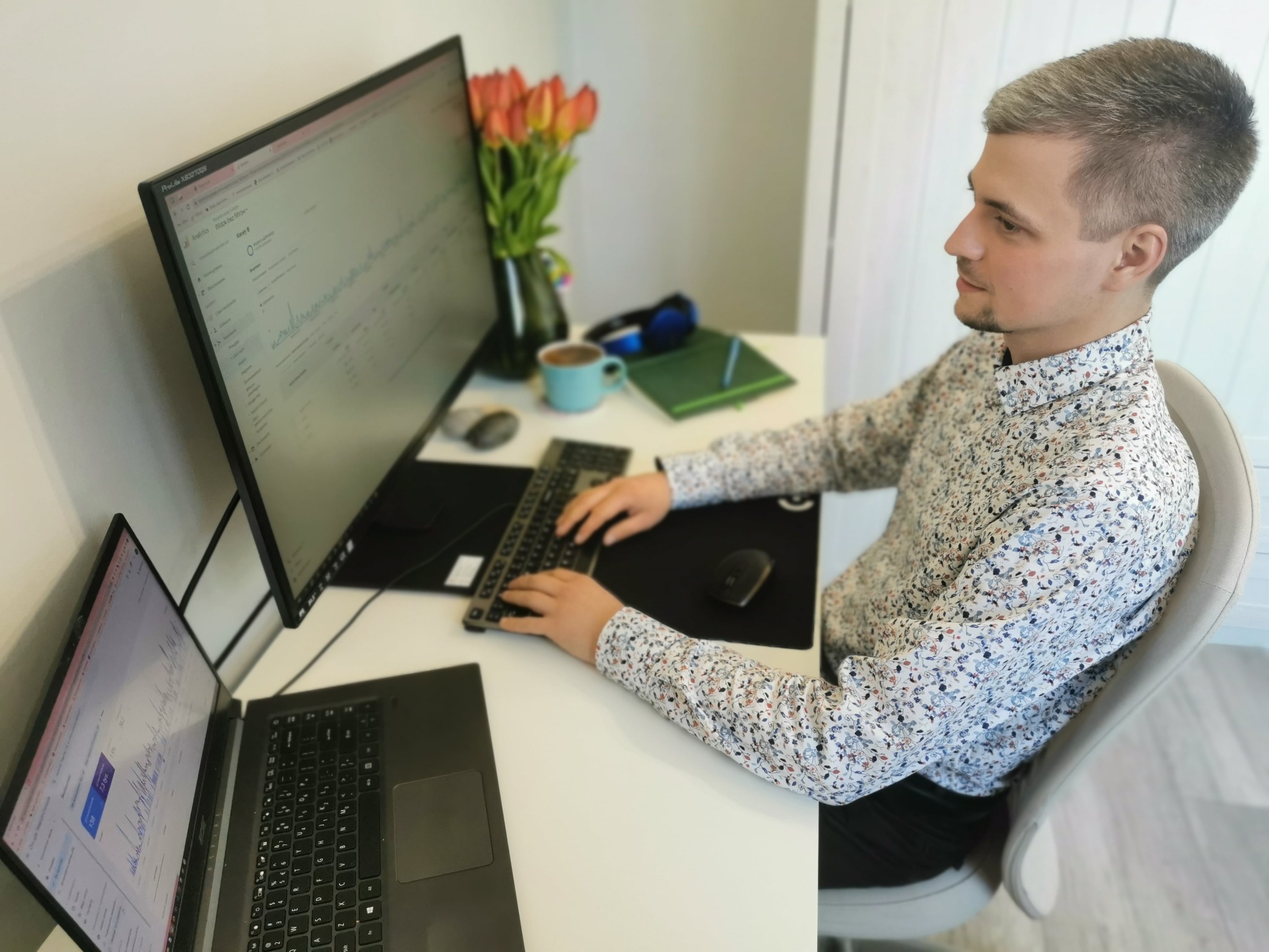 Mężczyzna w kwiecistej koszuli siedzi przy biurku z dwoma monitorami i laptopem, analizując wykresy, obok wazon z tulipanami i niebieski kubek.