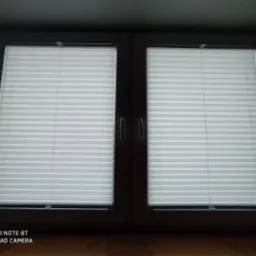 Dwuokienne okno z zamontowanymi, jasnymi plisami harmonijkowymi, widoczne uchwyty sterujące, ciemne tło pomieszczenia.