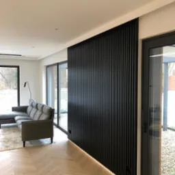 Nowoczesny salon z podświetlaną, pionową ścianką z czarnych paneli drewnianych, parkiet ułożony w jodełkę, szara sofa i duże okna z widokiem na ogród.