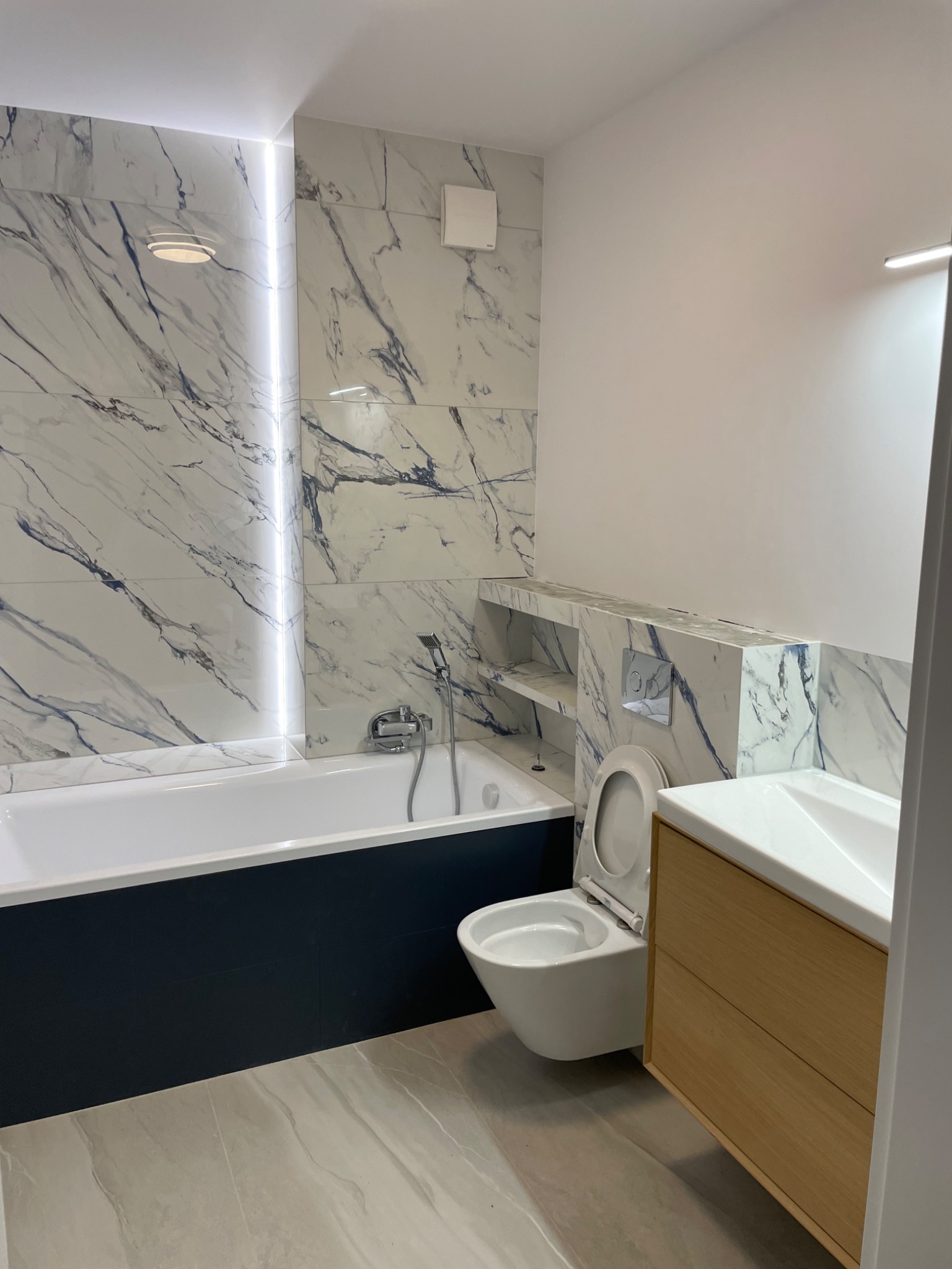 Nowoczesna łazienka z marmurowymi płytkami na ścianach, zabudowaną wanną w kolorze granatowym, podwieszaną toaletą i umywalką z drewnianą szafką, oświetleniem LED wzdłuż krawędzi płytek i wnęką...