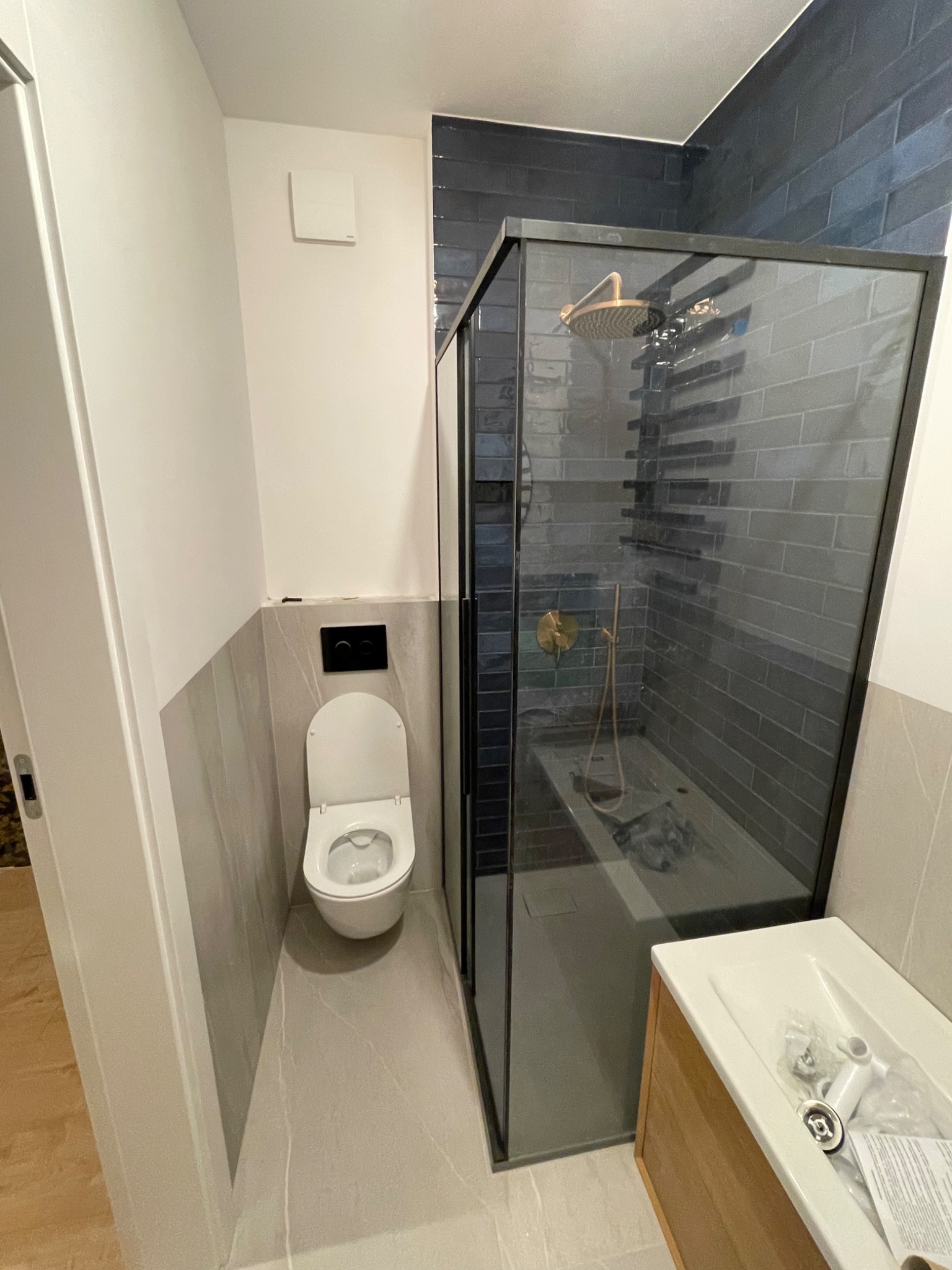 Nowoczesna łazienka z kabiną prysznicową typu walk-in z czarnymi ramami, płytkami w odcieniach szarości i złotą armaturą, widoczna toaleta i umywalka z elementami instalacyjnymi.