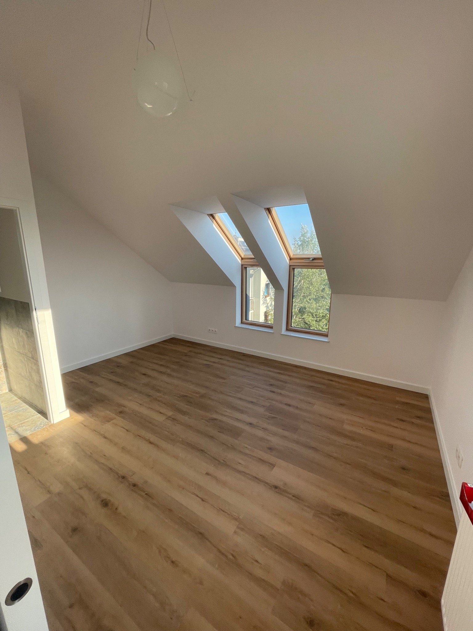 Poddasze z drewnopodobną podłogą, dwoma oknami dachowymi i białą, wiszącą lampą kulą. Widoczny fragment drzwi z szarą okładziną po lewej stronie.