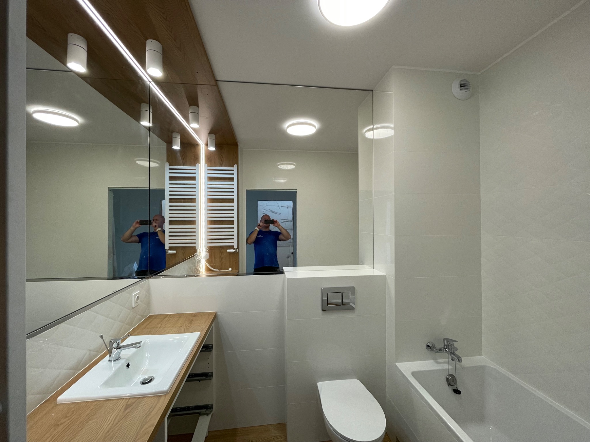 Nowoczesna łazienka z białymi płytkami, drewnianym blatem pod umywalką, dużym lustrem z oświetleniem LED i grzejnikiem drabinkowym, widoczna toaleta i wanna.