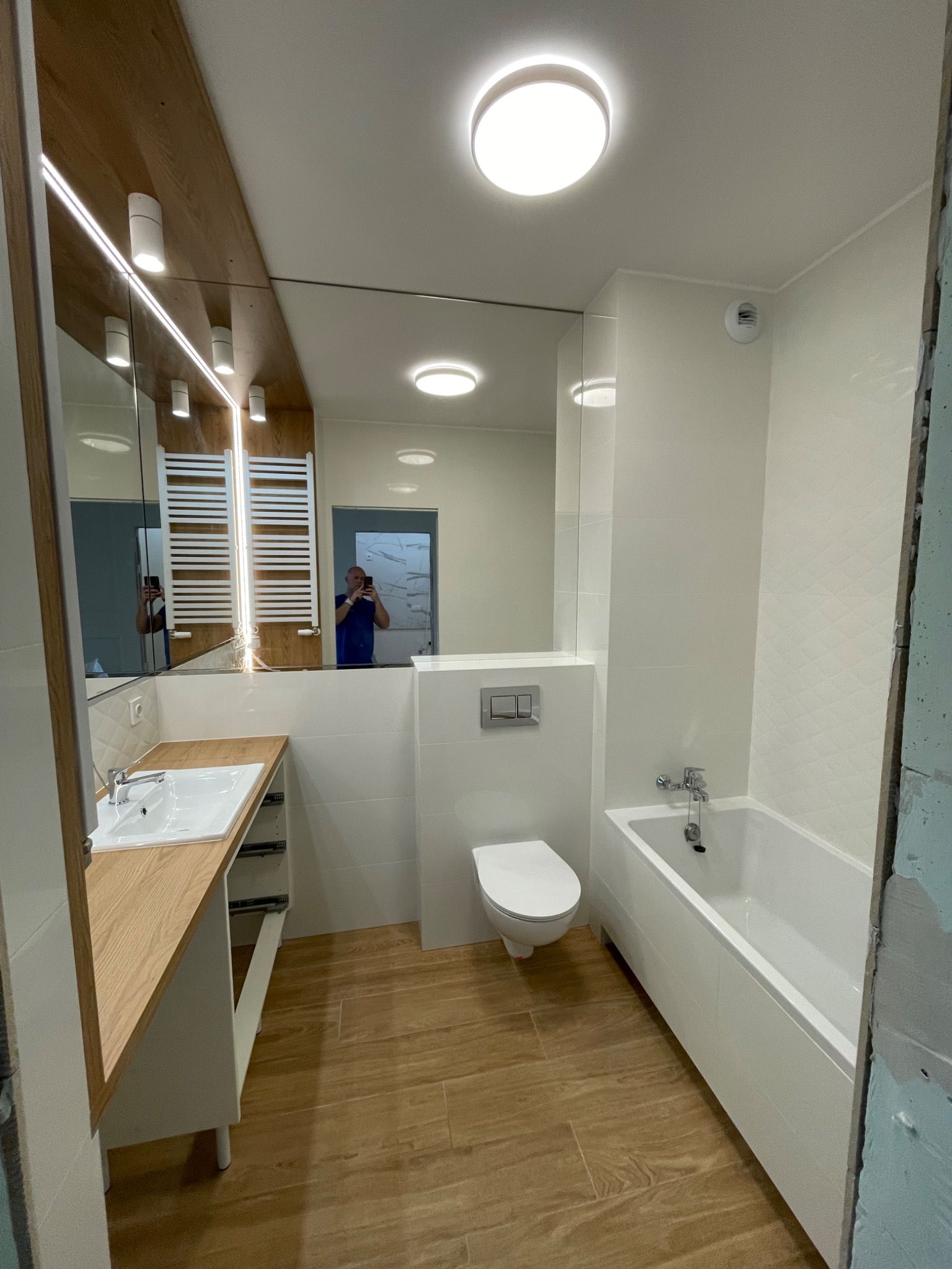 Nowoczesna łazienka z drewnianą podłogą, białymi płytkami z subtelnym wzorem rombów, umywalką z drewnianym blatem i dużym lustrem, w którym odbija się osoba robiąca zdjęcie.