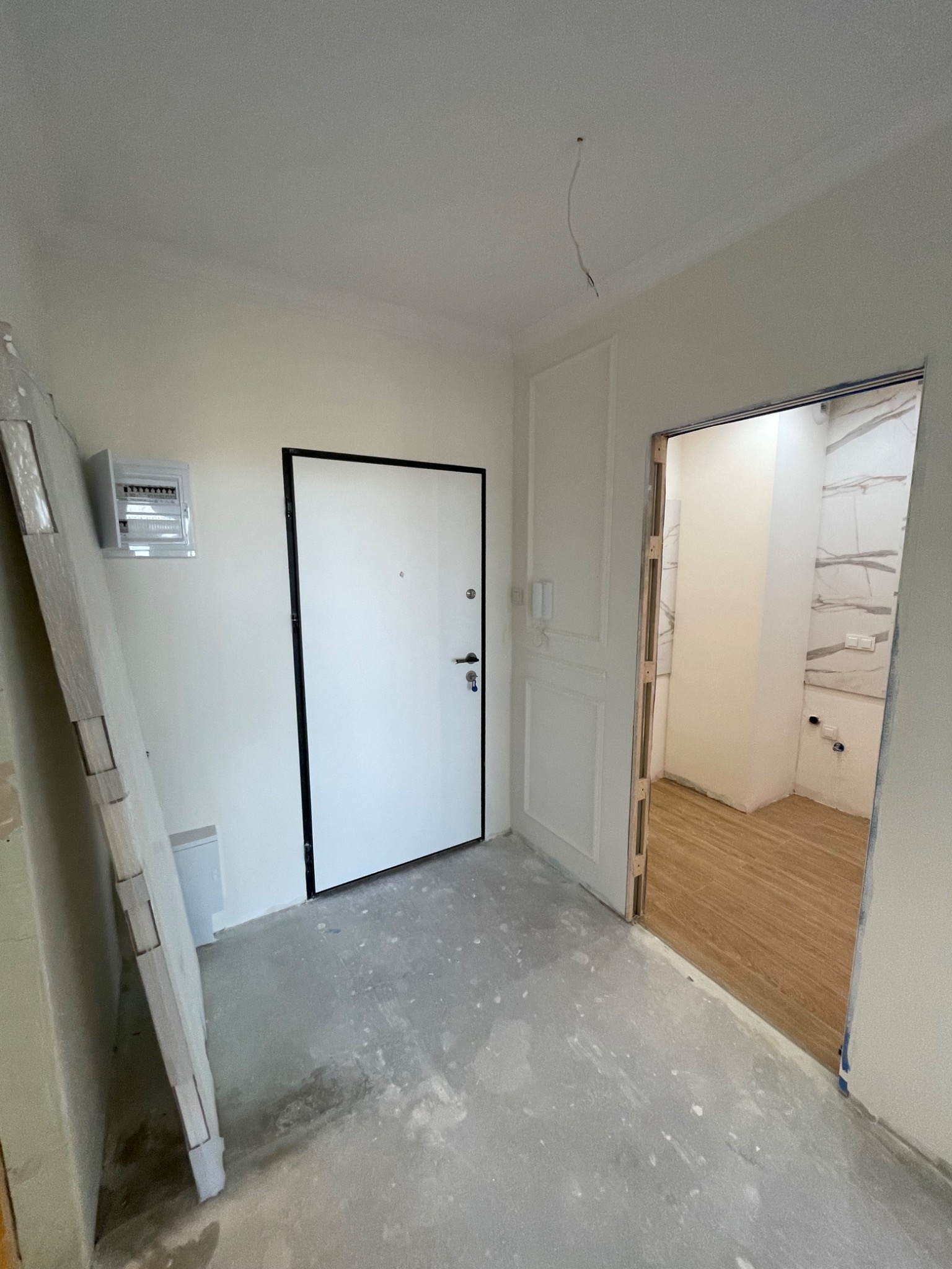 Wnętrze w trakcie prac w Pruszkowie: widok na białe drzwi z czarną ościeżnicą, surowy betonowy podkład, przygotowane do montażu panele przy ścianie i otwarte przejście do pomieszczenia...