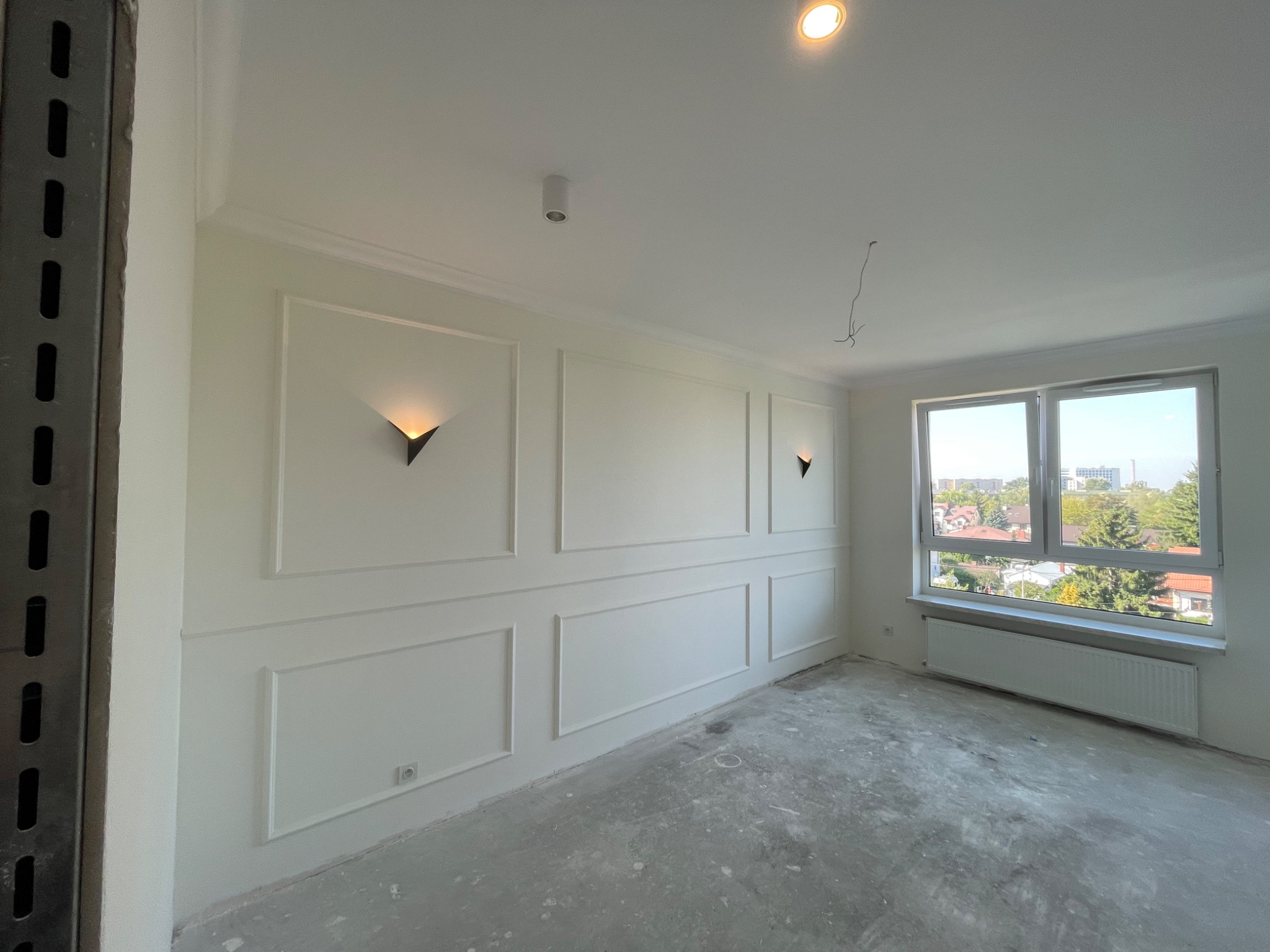Pusty pokój w trakcie remontu, ściany z białymi panelami dekoracyjnymi i nowoczesnymi lampami ściennymi, surowa podłoga przed położeniem parkietu, widok na miasto przez okno.