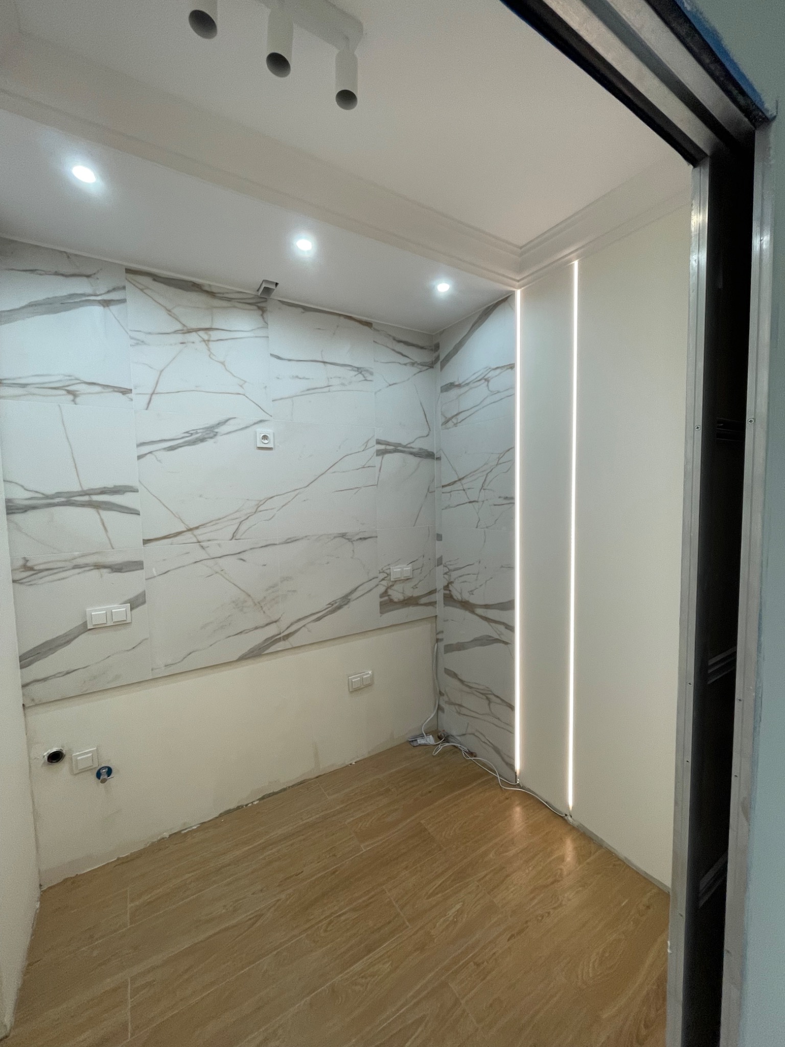 Wnętrze pomieszczenia z wykończoną podłogą imitującą drewno, ścianami z płytek imitujących marmur z widocznymi gniazdkami i włącznikami oraz pionowymi liniami świetlnymi na ścianie, widok zza...