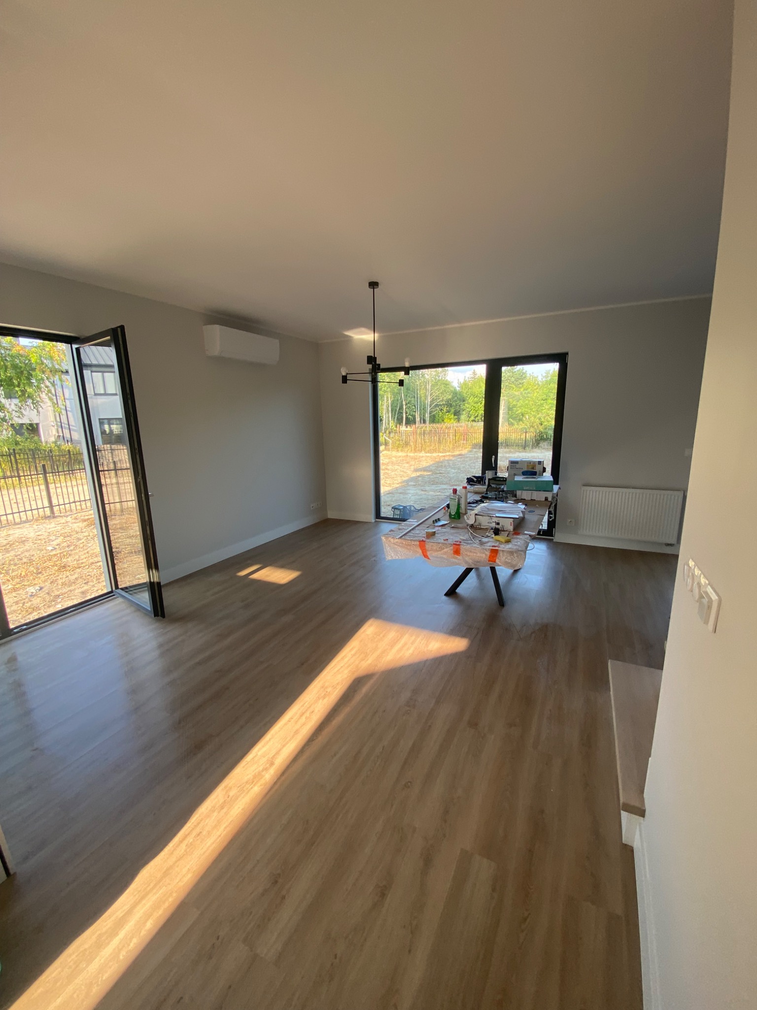 Jasne wnętrze domu w trakcie wykończenia, z drewnopodobną podłogą, dużymi oknami i stołem roboczym z narzędziami i materiałami budowlanymi, przygotowane do dalszych prac wykończeniowych.