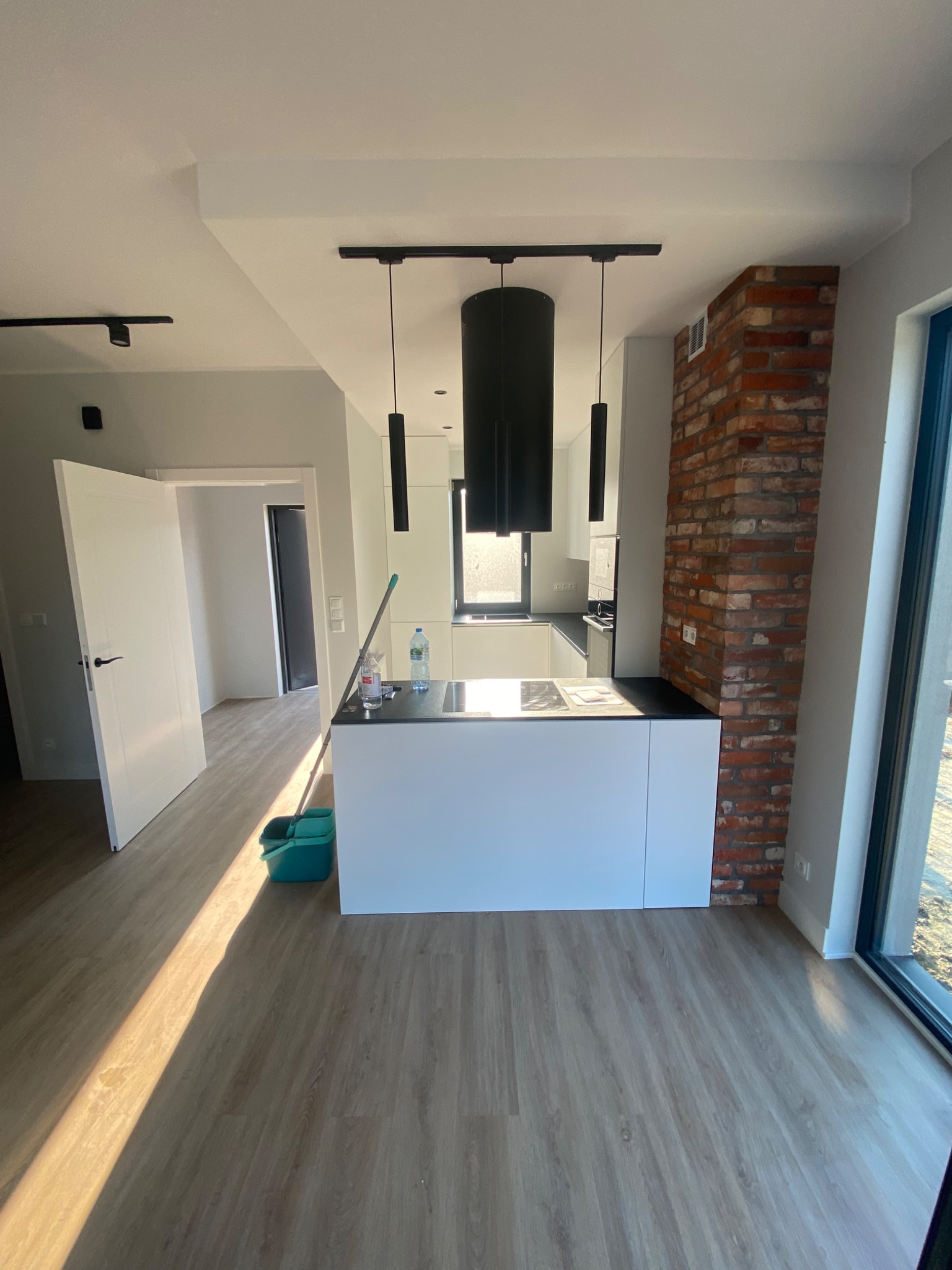 Nowoczesna kuchnia w trakcie wykańczania, z białymi meblami, ceglanym elementem dekoracyjnym, drewnopodobną podłogą, czarnymi lampami i widocznym sprzętem do mycia podłogi.