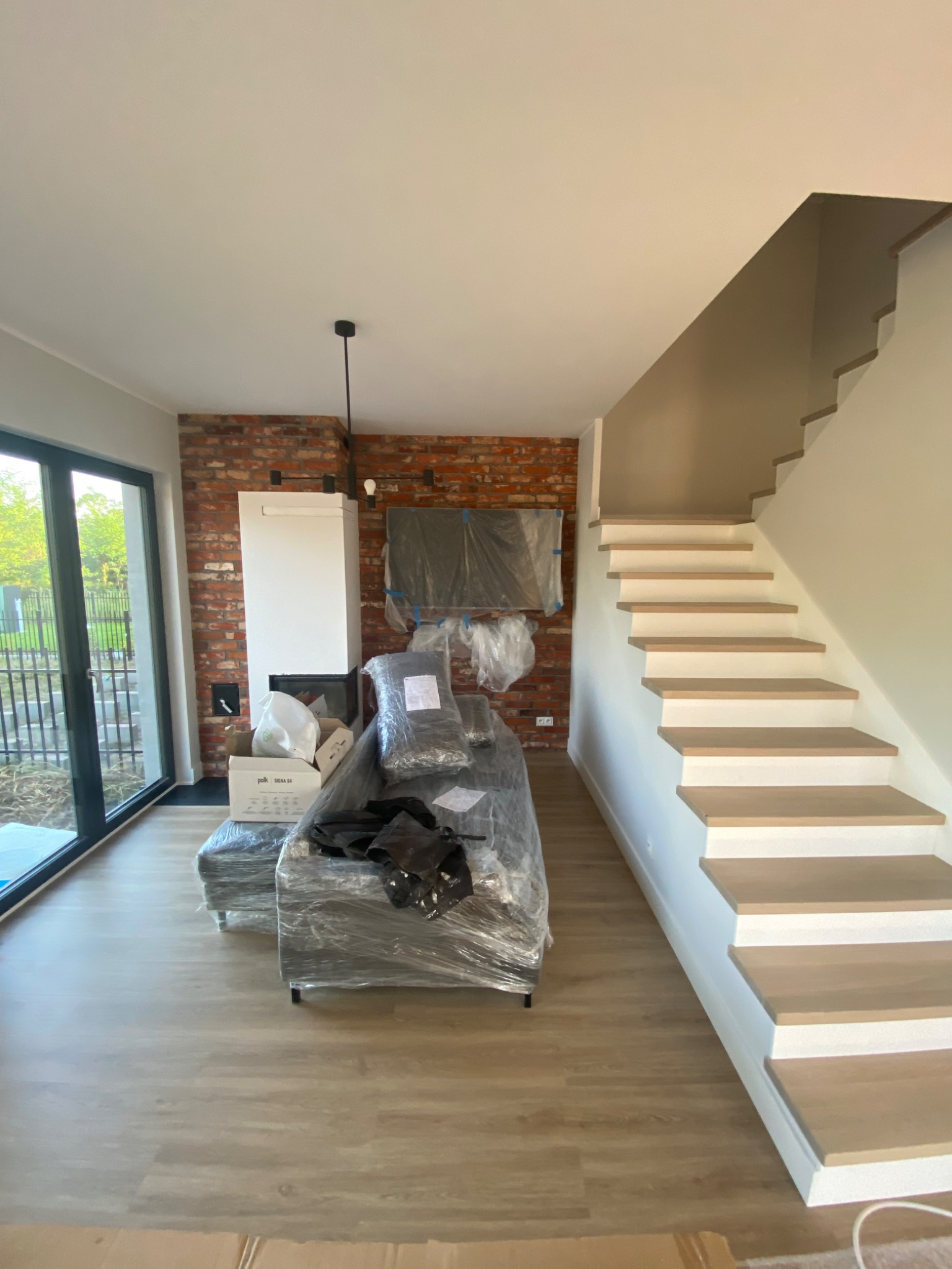 Nowe mieszkanie w trakcie urządzania: sofa i telewizor zabezpieczone folią, widoczny kominek, ceglana ściana, drewniane schody i panele podłogowe.