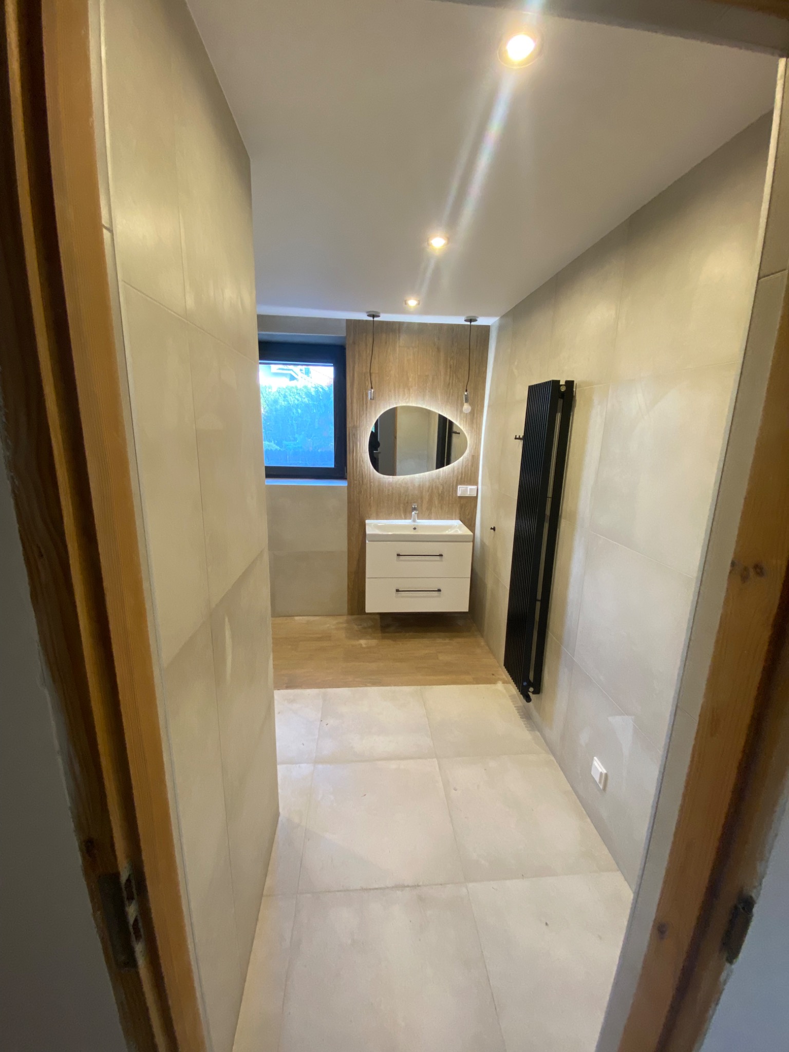 Nowoczesna łazienka z beżowymi płytkami, owalnym lustrem z podświetleniem LED, białą szafką pod umywalką i czarnym grzejnikiem drabinkowym, widoczna z perspektywy otwartych drzwi.