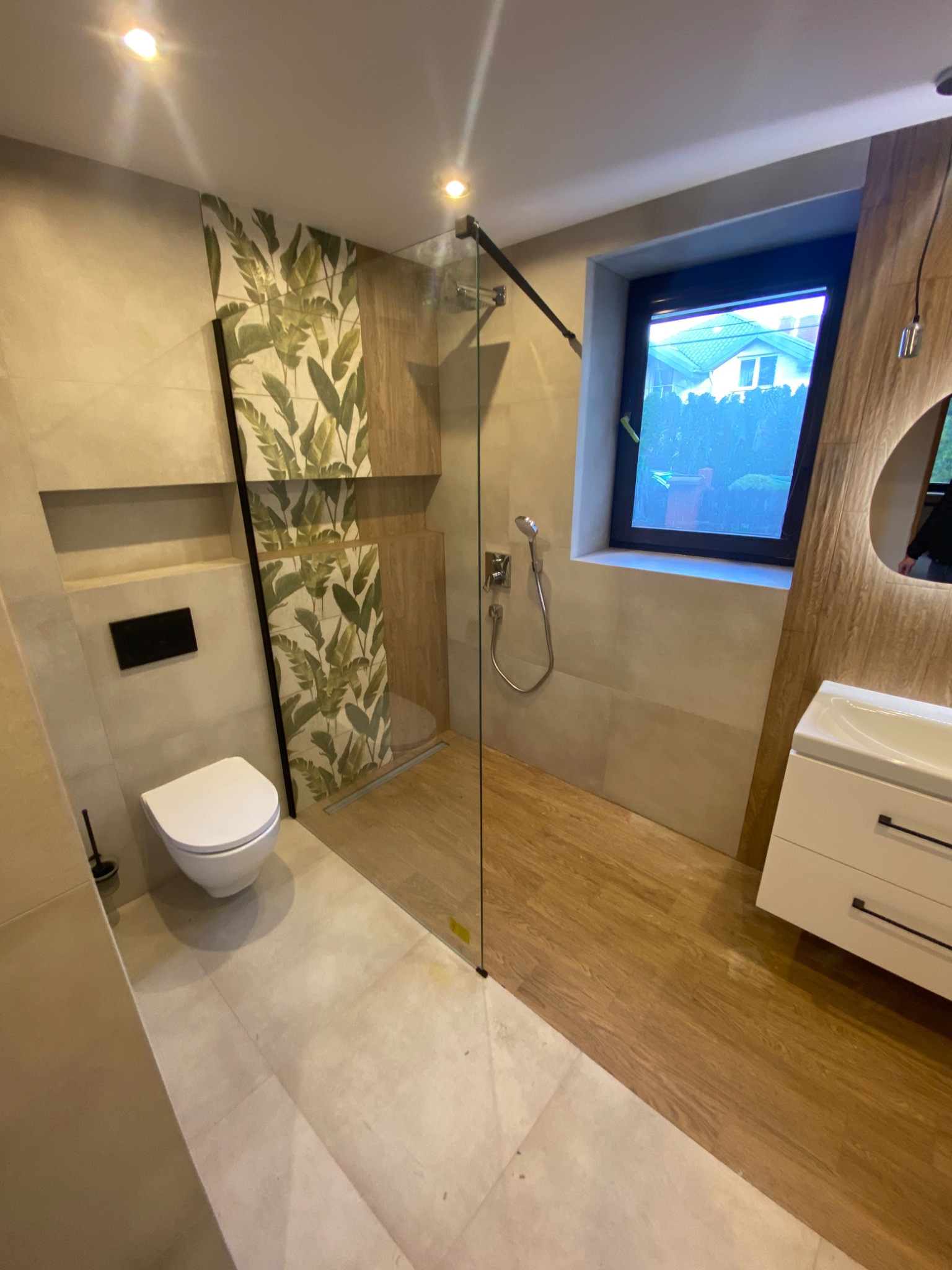 Nowoczesna łazienka z kabiną prysznicową typu walk-in, toaletą podwieszaną, umywalką z szafką i płytkami imitującymi drewno na podłodze w strefie prysznica, oraz akcentem w postaci płytek z motywem...