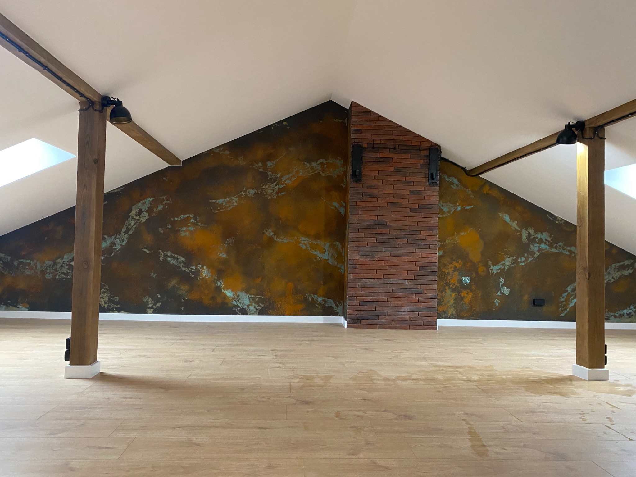 Poddasze po remoncie: drewniane belki konstrukcyjne z oświetleniem, ściana z efektem rdzy i fragment ściany wyłożony cegłą, jasna podłoga z paneli.