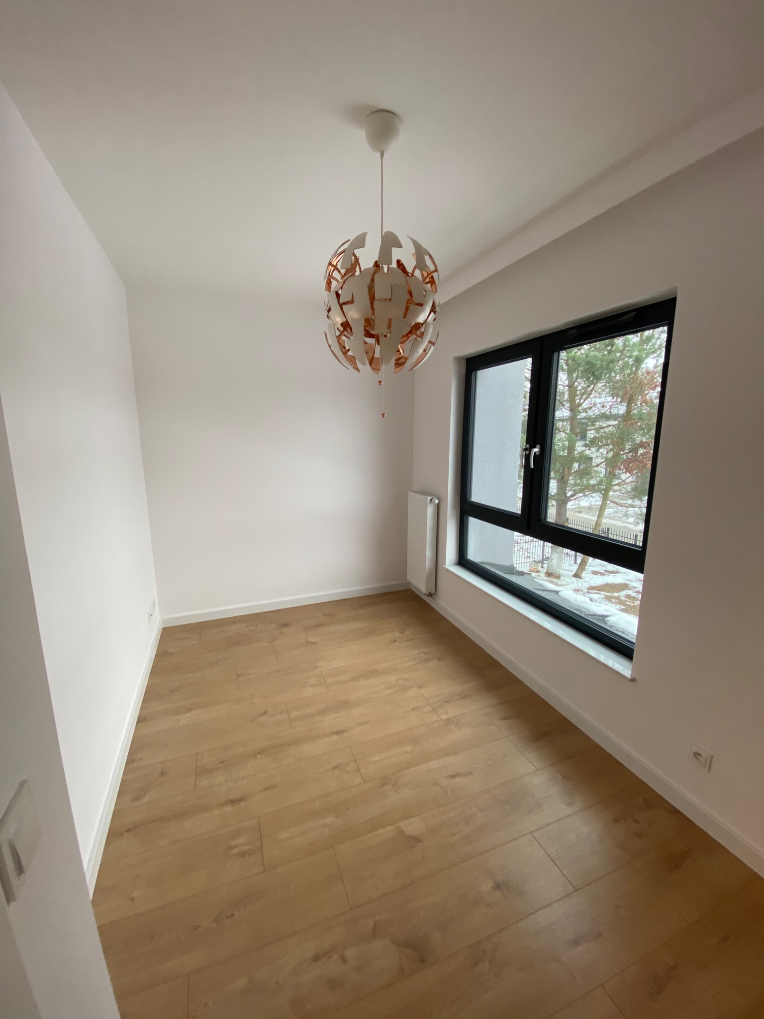 Puste, nowocześnie wykończone wnętrze z drewnianą podłogą, białymi ścianami i designerską lampą wiszącą, widok przez okno na drzewa pokryte śniegiem.