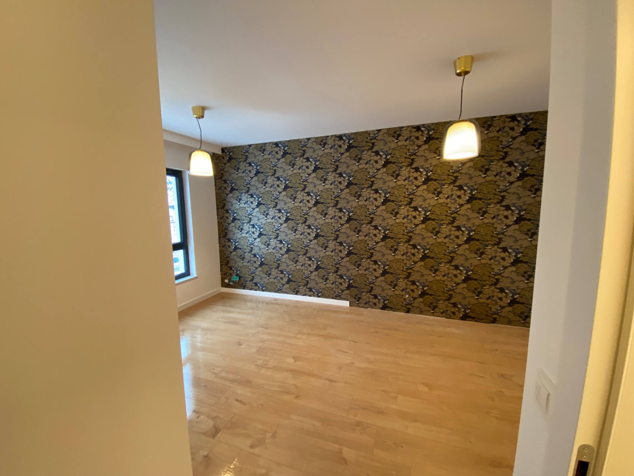 Pokój po remoncie: drewnopodobna podłoga, ściana w ciemną tapetę z roślinnym motywem, dwie wiszące lampy z ciepłym światłem, widok zza framugi drzwi.