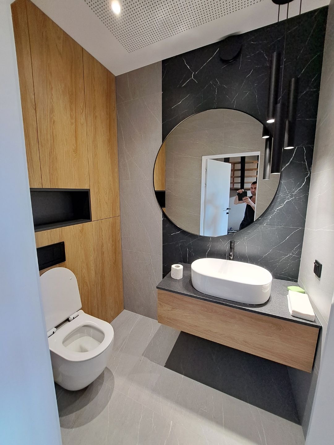 Nowoczesna łazienka z drewnianymi szafkami, okrągłym lustrem i czarną ścianą z marmurowym wzorem, widok na toaletę i umywalkę nablatową, oświetlenie punktowe.