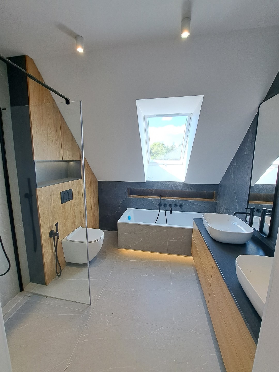 Nowoczesna łazienka na poddaszu z oknem dachowym, szarą armaturą i drewnianymi akcentami, oświetlona listwą LED pod wanną.